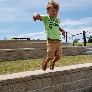 May include: A young boy wearing a green shirt with the text "JESUS LOVES ME" in black letters jumps in the air in front of a stone wall. The boy is wearing brown shorts and red sandals.
