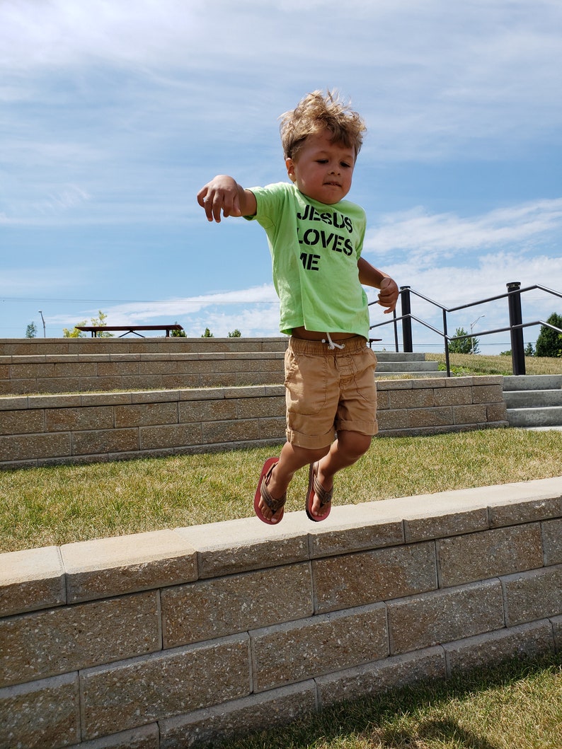 May include: A young boy wearing a green shirt with the text "JESUS LOVES ME" in black letters jumps in the air in front of a stone wall. The boy is wearing brown shorts and red sandals.