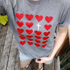May include: A gray t-shirt with a pattern of red hearts and a white cross in the center. The shirt has a rolled-up sleeve.