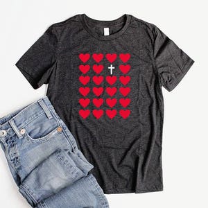 May include: A dark grey t-shirt with a pattern of red hearts and a white cross in the center. The hearts are arranged in a grid pattern.
