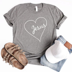 May include: A gray t-shirt with a white heart design that says "Jesus" inside the heart. The shirt is laid out on a white background with a pair of brown sandals, a white baseball cap, and blue jeans.