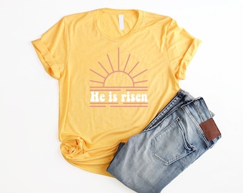 May include: A yellow t-shirt with a pink sun and the text "He is risen" in white. The shirt is folded and placed on top of a pair of blue jeans.
