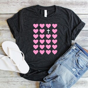 May include: A dark gray t-shirt with a pink heart design. The hearts are arranged in a grid with a white cross in the center.