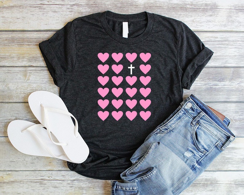 May include: A dark gray t-shirt with a pink heart design. The hearts are arranged in a grid with a white cross in the center.