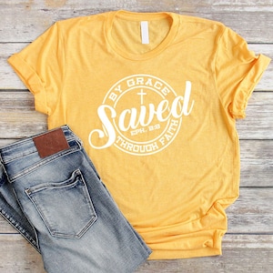 May include: A yellow t-shirt with a white circle graphic that says "Saved by Grace through Faith" with a cross and the scripture reference "Eph. 2:8".