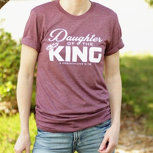 Op de afbeelding: Een maroon t-shirt met de tekst "Daughter of the King" en "2 Corinthians 6:18" in wit. De tekst is in een cursief schrift en heeft een doornenkroon boven het woord "King".