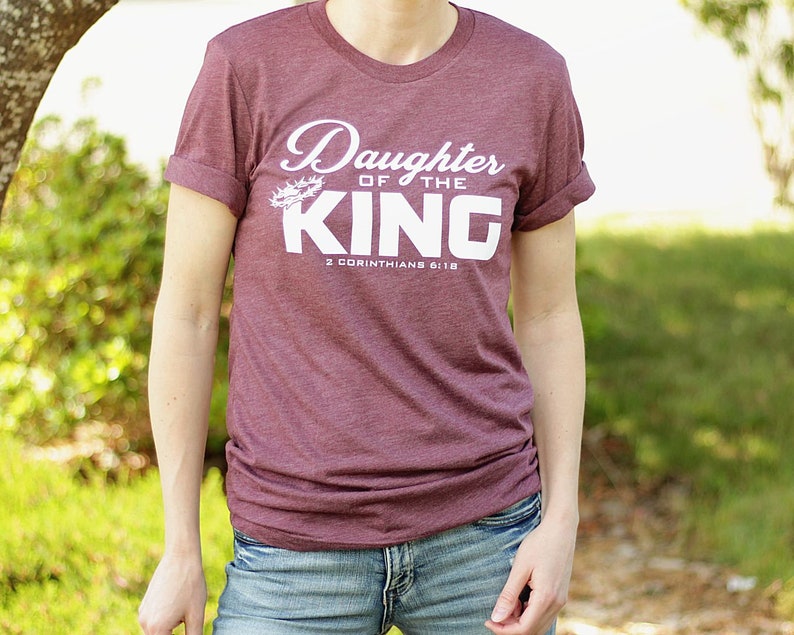 Op de afbeelding: Een maroon t-shirt met de tekst "Daughter of the King" en "2 Corinthians 6:18" in wit. De tekst is in een cursief schrift en heeft een doornenkroon boven het woord "King".