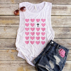 K&ouml;nnte beinhalten: Ein wei&szlig;es Tanktop mit einem Aufdruck von rosa Herzen und einem schwarzen Kreuz in der Mitte. Das Tanktop liegt auf einer Holzoberfl&auml;che mit einer Sonnenbrille und einer Jeans-Shorts.