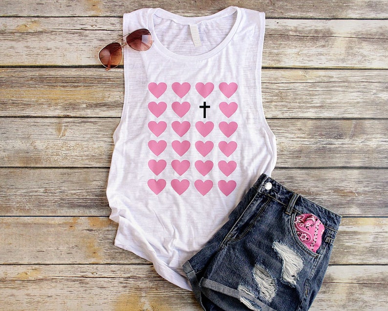 K&ouml;nnte beinhalten: Ein wei&szlig;es Tanktop mit einem Aufdruck von rosa Herzen und einem schwarzen Kreuz in der Mitte. Das Tanktop liegt auf einer Holzoberfl&auml;che mit einer Sonnenbrille und einer Jeans-Shorts.
