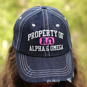 May include: A blue denim trucker hat with a white mesh back. The hat has white stitching and a pink and white logo that says "PROPERTY OF AΩ ALPHA & OMEGA".