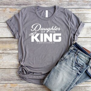 Op de afbeelding: Grijs T-shirt met de tekst "Daughter of the King" en de bijbelverwijzing "2 Korinthe 6:18".