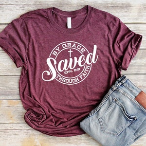 May include: A maroon t-shirt with a white circle graphic that says "Saved by Grace through Faith" with a cross and the scripture reference "Eph. 2:8".