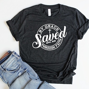 May include: A dark grey t-shirt with a white circle graphic that says "Saved by Grace through Faith" with a cross and the scripture reference "Eph. 2:8".