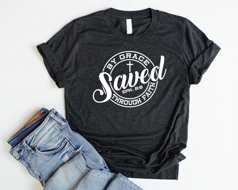 May include: A dark grey t-shirt with a white circle graphic that says "Saved by Grace through Faith" with a cross and the scripture reference "Eph. 2:8".