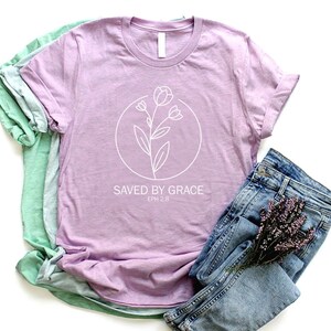 K&ouml;nnte beinhalten: Ein lavendelfarbenes T-Shirt mit einer wei&szlig;en Umrandung einer Blume und dem Text "SAVED BY GRACE EPH 2:8". Das Shirt ist gefaltet und wird mit einer blauen Jeans und einem Lavendelzweig pr&auml;sentiert.