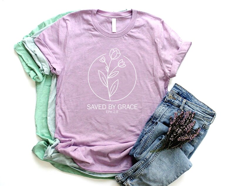 K&ouml;nnte beinhalten: Ein lavendelfarbenes T-Shirt mit einer wei&szlig;en Umrandung einer Blume und dem Text "SAVED BY GRACE EPH 2:8". Das Shirt ist gefaltet und wird mit einer blauen Jeans und einem Lavendelzweig pr&auml;sentiert.