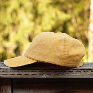 May include: A yellow baseball cap with a slightly distressed look. The cap is sitting on a wooden surface.