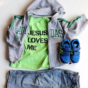 May include: A gray hooded sweatshirt with green stripes and the word "DAS" on the right side. A green t-shirt with the words "JESUS LOVES ME" in black letters is worn underneath. Blue jeans and blue sneakers with green accents are also included.
