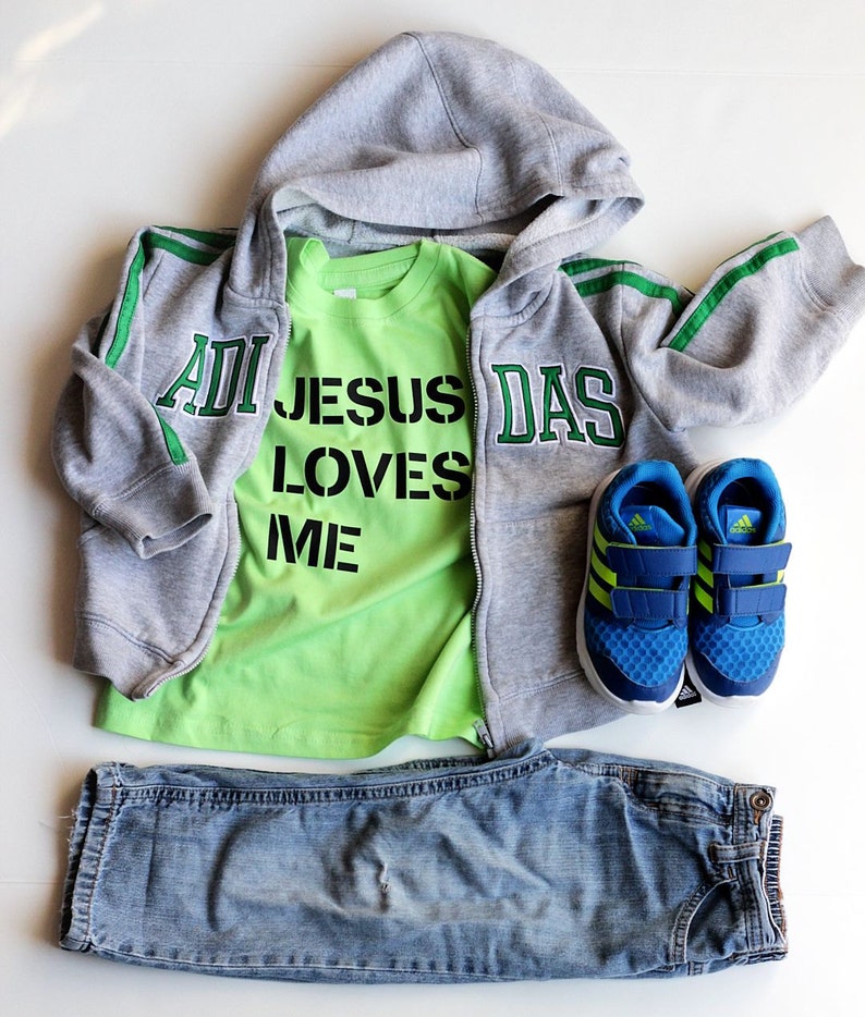May include: A gray hooded sweatshirt with green stripes and the word "DAS" on the right side. A green t-shirt with the words "JESUS LOVES ME" in black letters is worn underneath. Blue jeans and blue sneakers with green accents are also included.