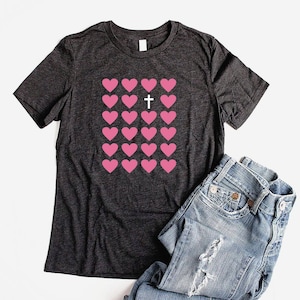May include: A dark gray t-shirt with a pink heart design and a white cross in the center of the hearts.
