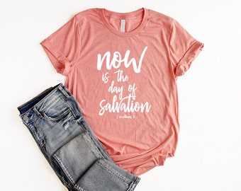 May include: A coral pink t-shirt with the text "NOW is the day of SALVATION" in white lettering. The text is in a handwritten font. The shirt is made of a soft, comfortable material and has a relaxed fit. The shirt is shown on a white background.