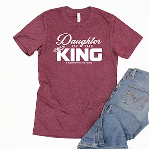 Op de afbeelding: Bordeauxkleurig heather t-shirt met witte tekst die "Daughter of the King" en "2 Corinthians 6:18" zegt.