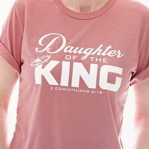 Op de afbeelding: Een roze T-shirt met de tekst "Daughter of the King" in wit met een doornenkroon-afbeelding en de Bijbelverwijzing "2 Corinthians 6:18".