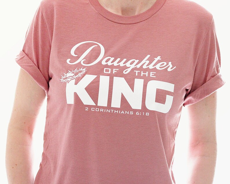 Op de afbeelding: Een roze T-shirt met de tekst "Daughter of the King" in wit met een doornenkroon-afbeelding en de Bijbelverwijzing "2 Corinthians 6:18".