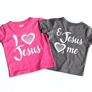 May include: Two t-shirts with a heart and the text "I love Jesus" and "& Jesus love me". The shirt on the left is pink and the shirt on the right is gray.