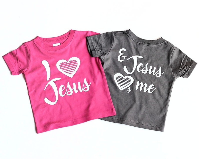 May include: Two t-shirts with a heart and the text "I love Jesus" and "& Jesus love me". The shirt on the left is pink and the shirt on the right is gray.