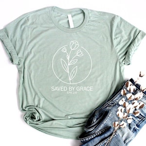 K&ouml;nnte beinhalten: Ein hellgr&uuml;nes T-Shirt mit einem wei&szlig;en Blumenmuster und dem Text "SAVED BY GRACE EPH 2:8".