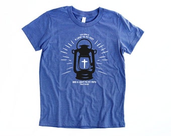 May include: A blue t-shirt with a white graphic of a lantern with a cross on it. The text on the shirt reads "Your word is a lamp to my feet and a light to my path Psalm 119:105".