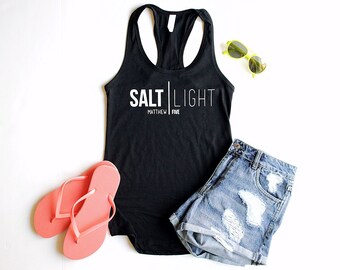May include: Black tank top with the text "SALT LIGHT" and "MATTHEW FIVE" in white. The tank top is paired with denim shorts and flip flops.