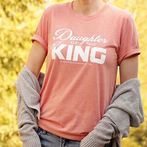 Op de afbeelding: Een koraalroze T-shirt met de tekst "Daughter of the King" en "2 Corinthians 6:18" in wit. Het shirt heeft een losse pasvorm en opgerolde mouwen.