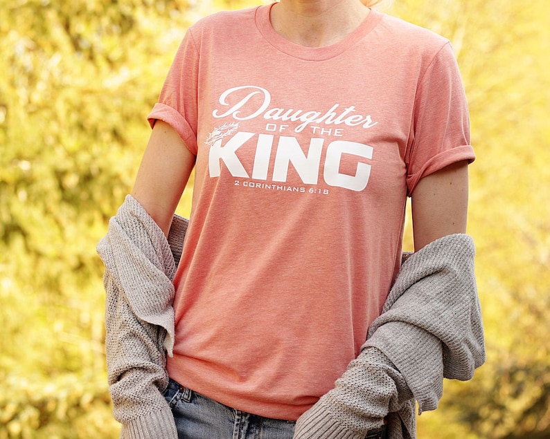 Op de afbeelding: Een koraalroze T-shirt met de tekst "Daughter of the King" en "2 Corinthians 6:18" in wit. Het shirt heeft een losse pasvorm en opgerolde mouwen.