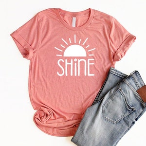 May include: A coral pink t-shirt with a white graphic of a sun with the word "shine" underneath.