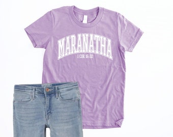 May include: A lavender t-shirt with the word "Maranatha" in white text with a white outline. The text is in an arch shape with the words "1 Cor 16-22" underneath. The t-shirt is paired with blue jeans.