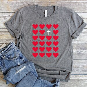 May include: A gray t-shirt with a design of red hearts and a white cross in the center. The hearts are arranged in a grid pattern.
