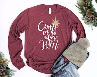 May include: A maroon long-sleeved t-shirt with the text "Come let us adore Him" in white lettering. A gold star is above the text. The shirt is laying on a pair of blue jeans and a beige beanie with a white pom-pom.