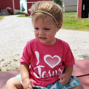 May include: A young child wearing a pink t-shirt with a white heart and the words "I Love Jesus" printed on it. The child is sitting on a wooden bench.