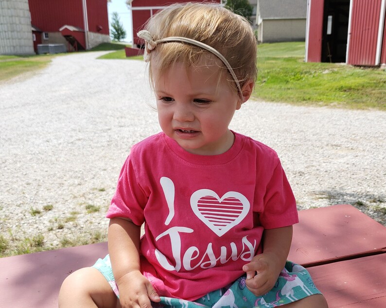 May include: A young child wearing a pink t-shirt with a white heart and the words "I Love Jesus" printed on it. The child is sitting on a wooden bench.