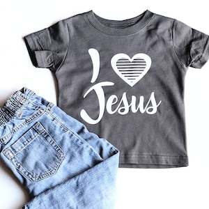 May include: A gray short-sleeved t-shirt with a white heart and the text "I love Jesus" printed on it. The shirt is paired with blue jeans.