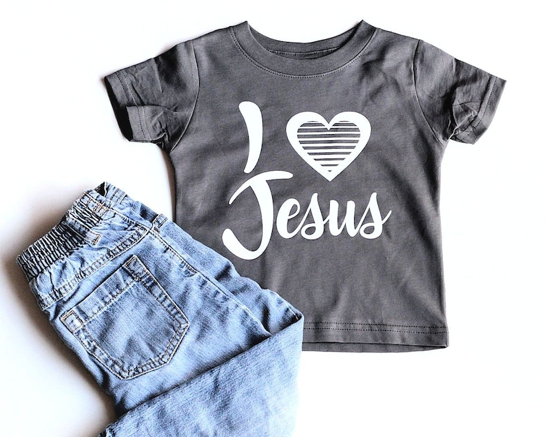 May include: A gray short-sleeved t-shirt with a white heart and the text "I love Jesus" printed on it. The shirt is paired with blue jeans.