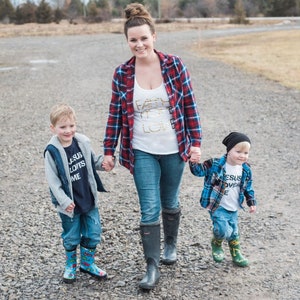 May include: A woman in a red and black plaid shirt and blue jeans walks with two young boys. The woman is wearing a white tank top with the words "Faith Hope Love" in gold glitter. The boys are wearing blue and white plaid shirts and blue jeans. The boy on the left is wearing a black vest and a blue shirt with the words "Jesus Loves Me" in white. The boy on the right is wearing a black beanie and a white shirt with the words "Jesus Loves Me" in black. All three are wearing rain boots.