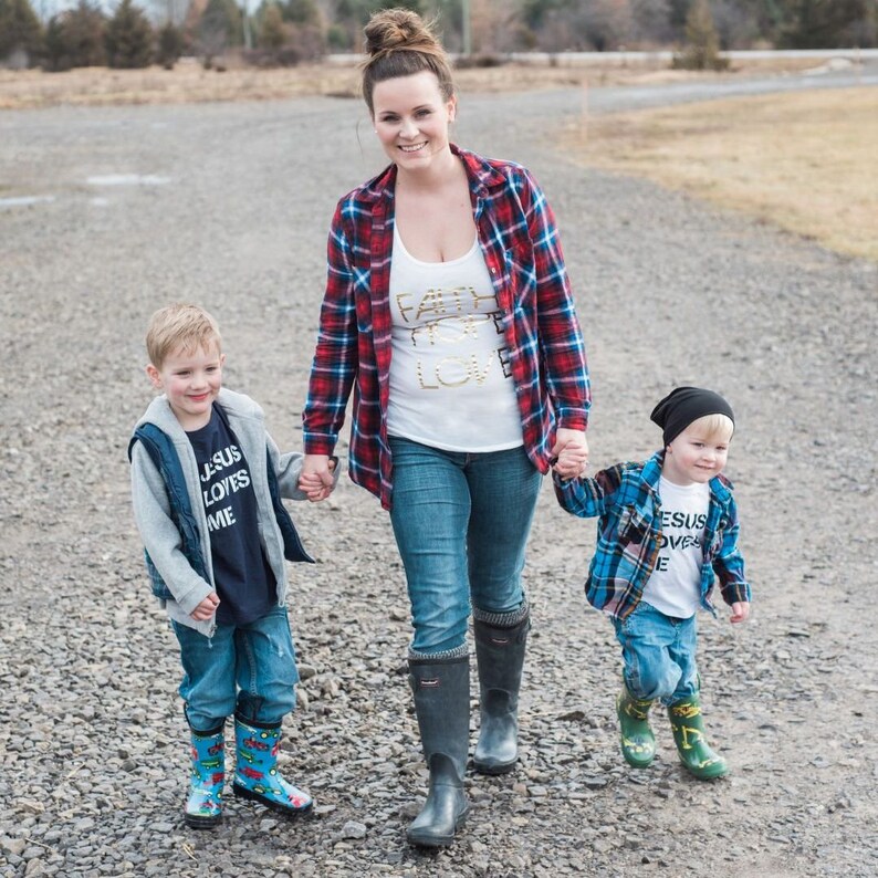 May include: A woman in a red and black plaid shirt and blue jeans walks with two young boys. The woman is wearing a white tank top with the words "Faith Hope Love" in gold glitter. The boys are wearing blue and white plaid shirts and blue jeans. The boy on the left is wearing a black vest and a blue shirt with the words "Jesus Loves Me" in white. The boy on the right is wearing a black beanie and a white shirt with the words "Jesus Loves Me" in black. All three are wearing rain boots.