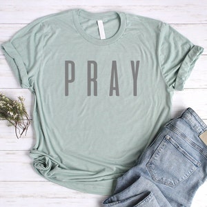 May include: A light green t-shirt with the word "PRAY" printed in gray on the front. The shirt is laid flat on a white wooden surface with a pair of blue jeans.