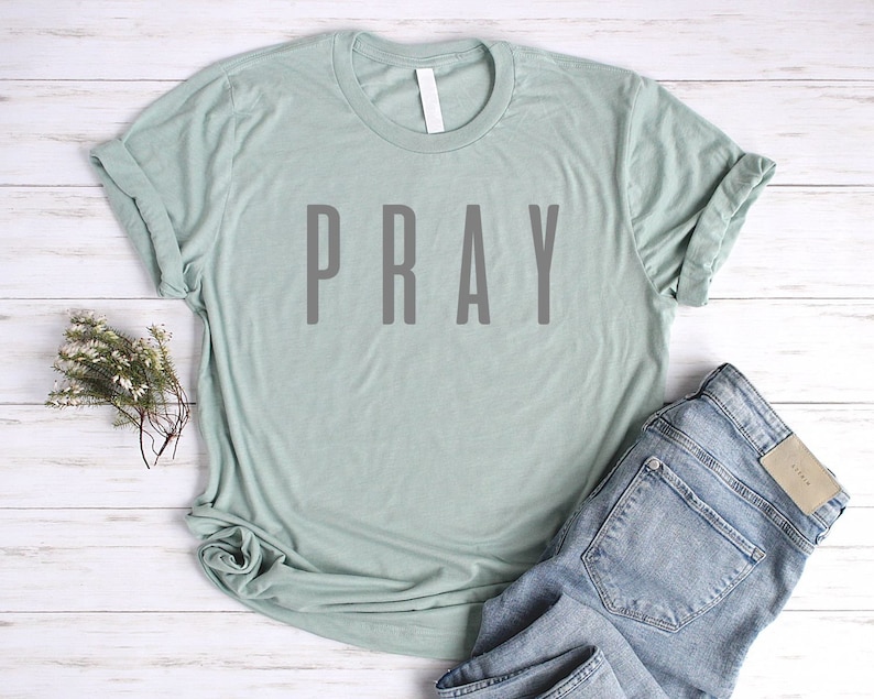 May include: A light green t-shirt with the word "PRAY" printed in gray on the front. The shirt is laid flat on a white wooden surface with a pair of blue jeans.