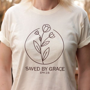 K&ouml;nnte beinhalten: Ein cremefarbenes T-Shirt mit einer braunen Linienzeichnung einer Blume in einem Kreis. Der Text "SAVED BY GRACE" und "EPH 2:8" ist unter der Blume aufgedruckt.
