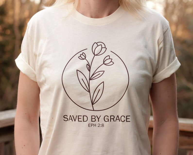 K&ouml;nnte beinhalten: Ein cremefarbenes T-Shirt mit einer braunen Linienzeichnung einer Blume in einem Kreis. Der Text "SAVED BY GRACE" und "EPH 2:8" ist unter der Blume aufgedruckt.