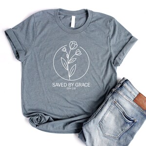 K&ouml;nnte beinhalten: Ein hellblaues T-Shirt mit einem wei&szlig;en Blumenmuster und dem Text "SAVED BY GRACE EPH 2:8".
